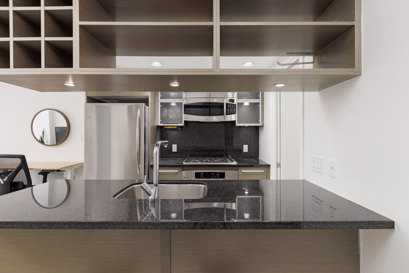 Modern kitchen with black granite countertops, a sink, stainless steel appliances, open shelving, and office area visible in the background.