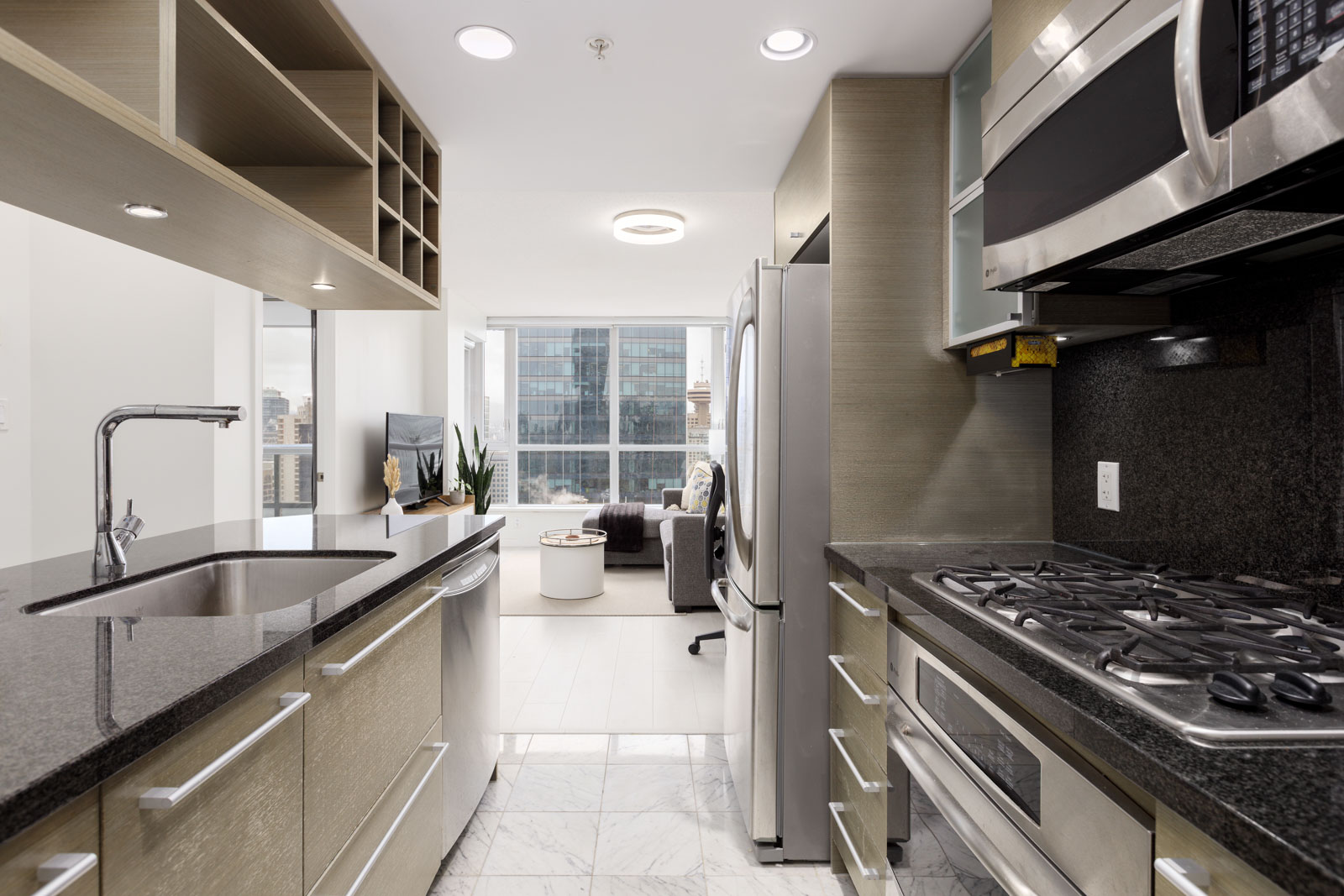 Modern kitchen with stainless steel appliances, gas stove, open shelving, dark countertops, and view of living area with large windows overlooking city buildings.