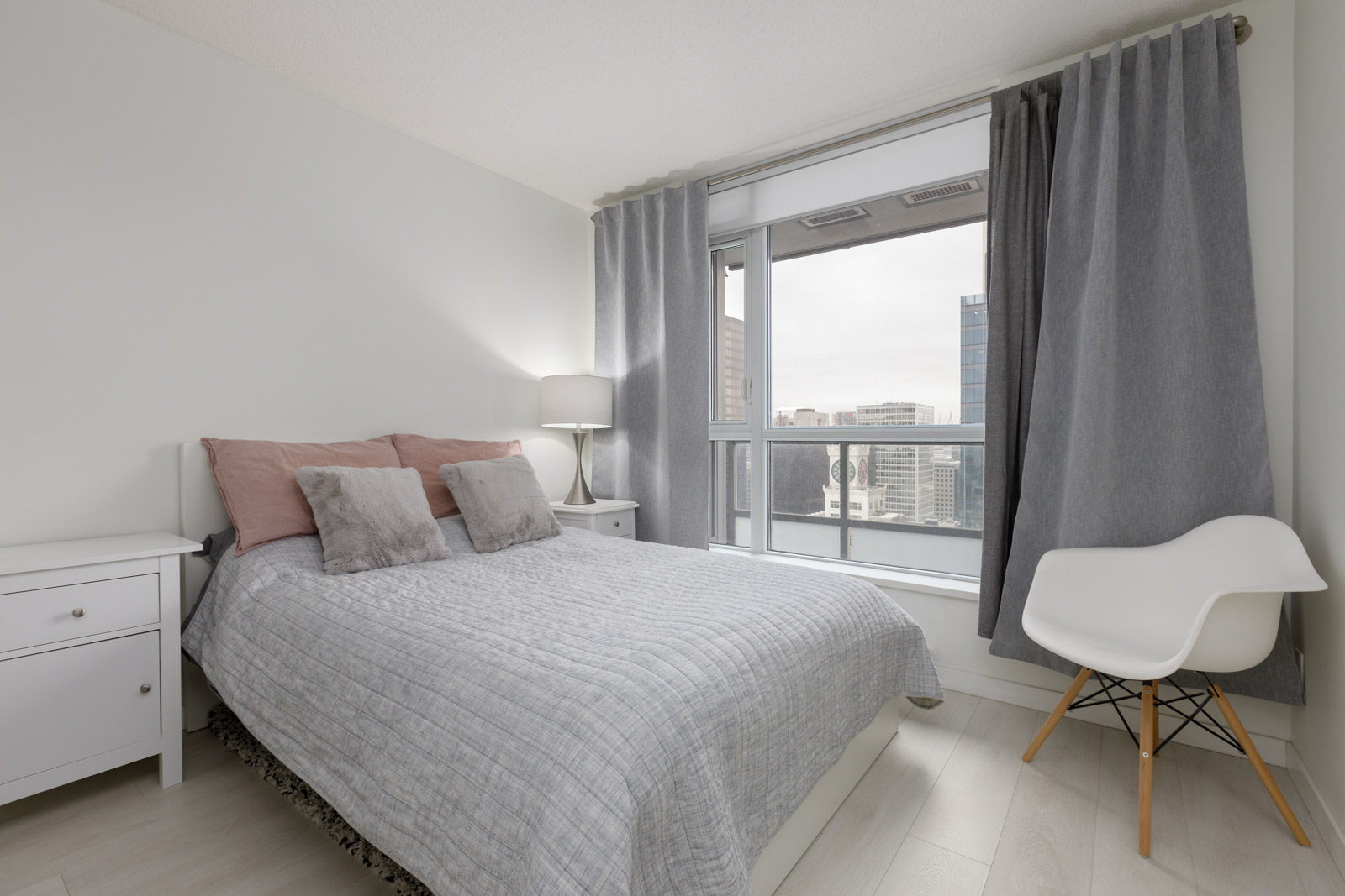 A modern bedroom with a neatly made bed, two bedside tables, a lamp, gray curtains, a white chair, and a large window overlooking city buildings.