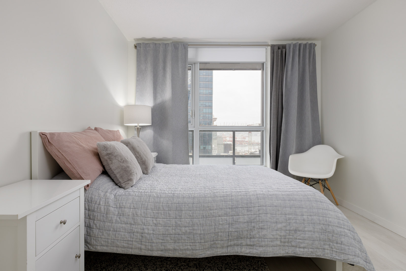 A modern, minimalist bedroom with a neatly made bed, two-tone pillows, a white nightstand, an accent chair, and large window with gray curtains overlooking city buildings.