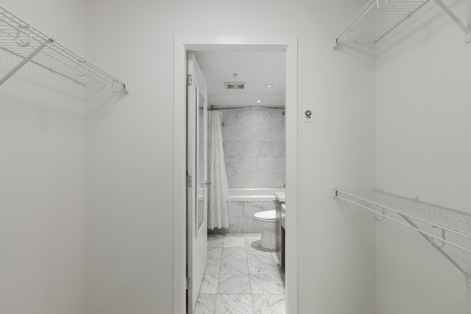 White walk-in closet with wire shelving on both sides, leading to a bathroom with marble flooring and walls, visible toilet, bathtub, and shower curtain.