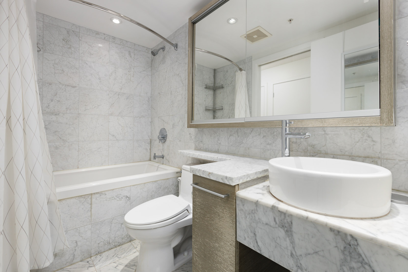 Modern bathroom with marble walls and counters, a white vessel sink, a toilet, a large mirror, and a bathtub with a shower curtain.