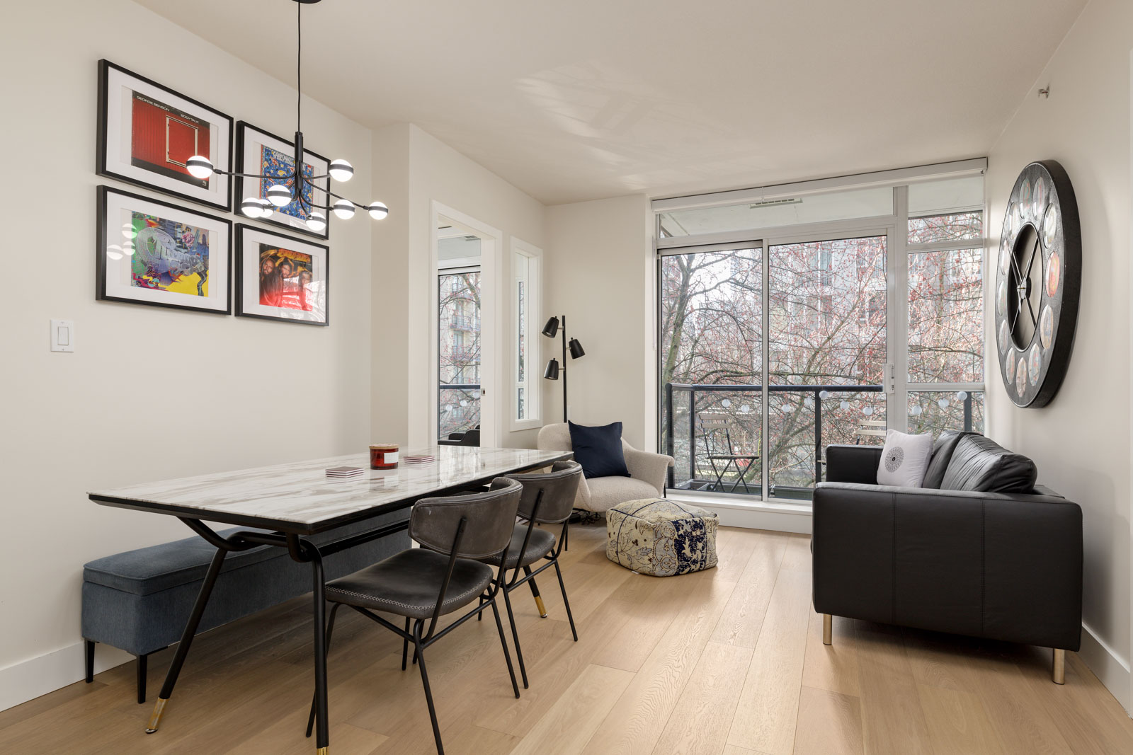 Modern dining and living area with a marble table, black chairs, a black sofa, large windows, light wood flooring, wall art, and a round wall clock.