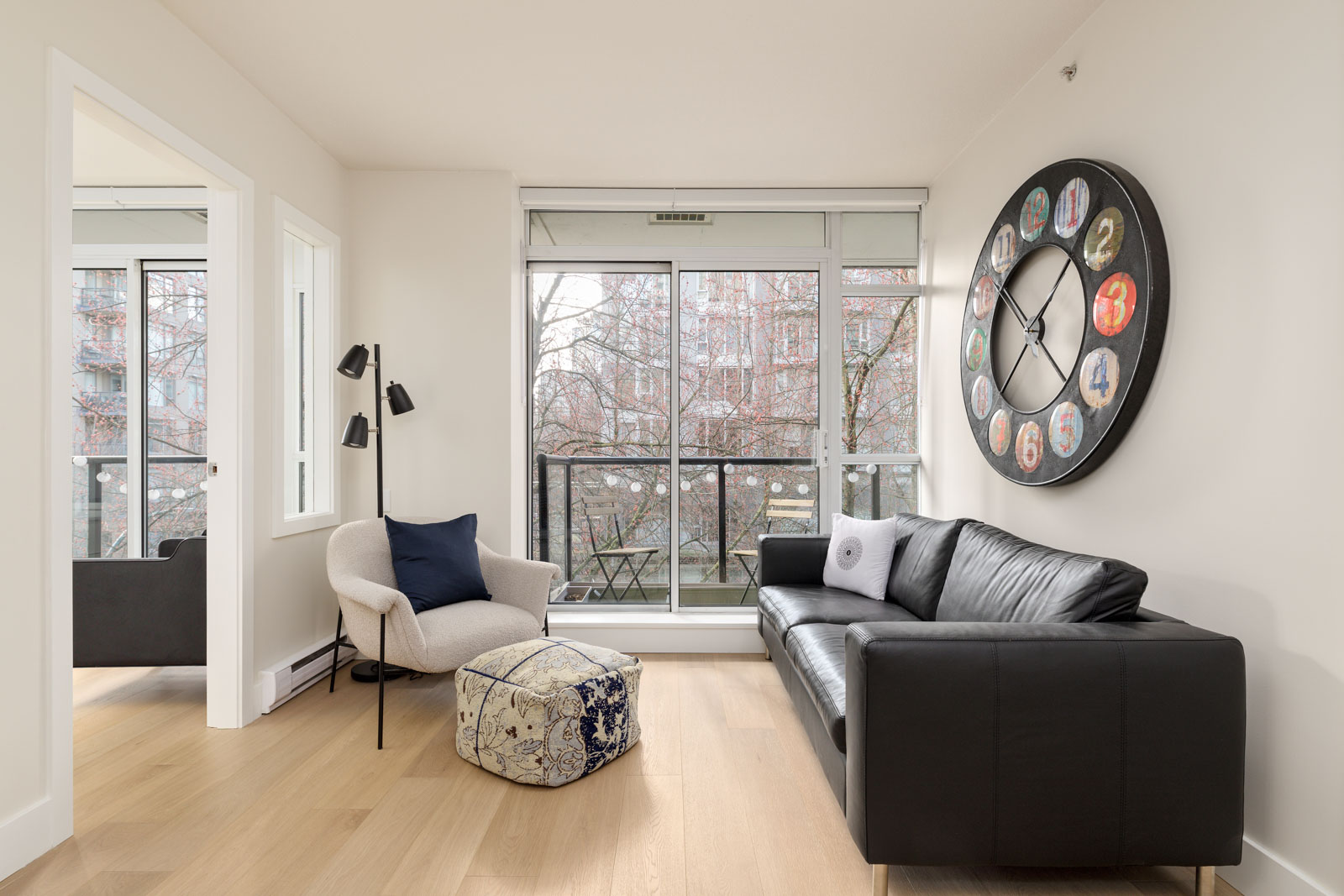 Modern living room with a black leather sofa, armchair, patterned pouf, large wall clock, floor lamp, and a balcony visible through glass doors.
