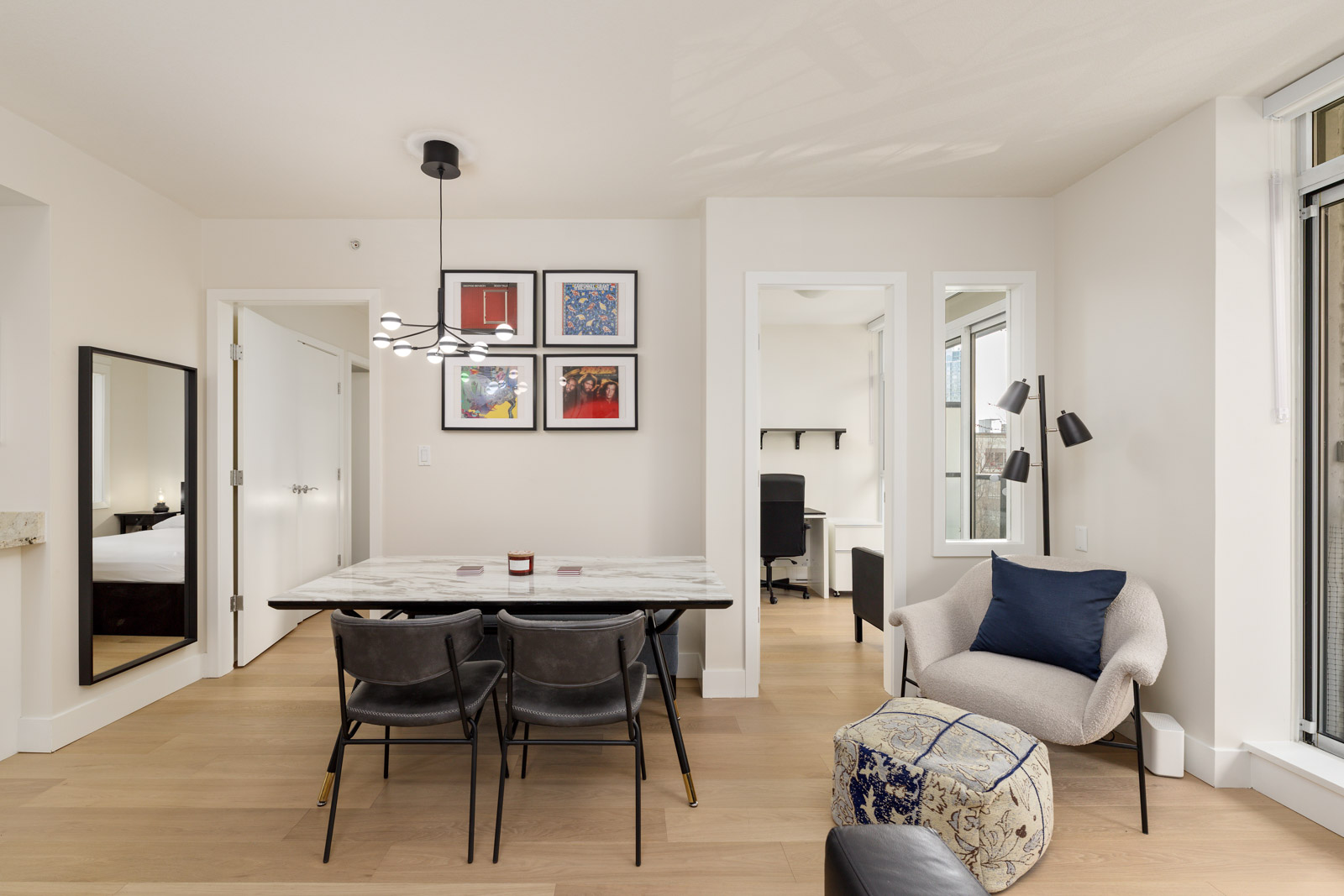 Modern dining and living area with a marble table, four chairs, armchair, pouf, wall art, and a small home office visible through glass panels.