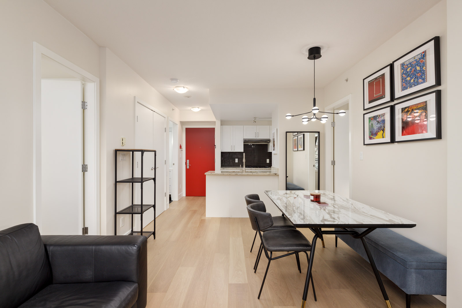 Modern apartment interior with a marble dining table, four black chairs, black couch, open kitchen, light wood flooring, and framed art on the wall.