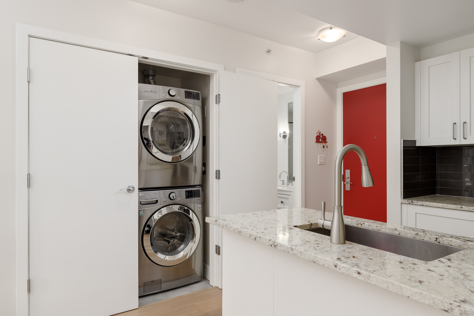A modern laundry area with stacked washer and dryer in a closet next to a kitchen with granite countertops and a red entry door.