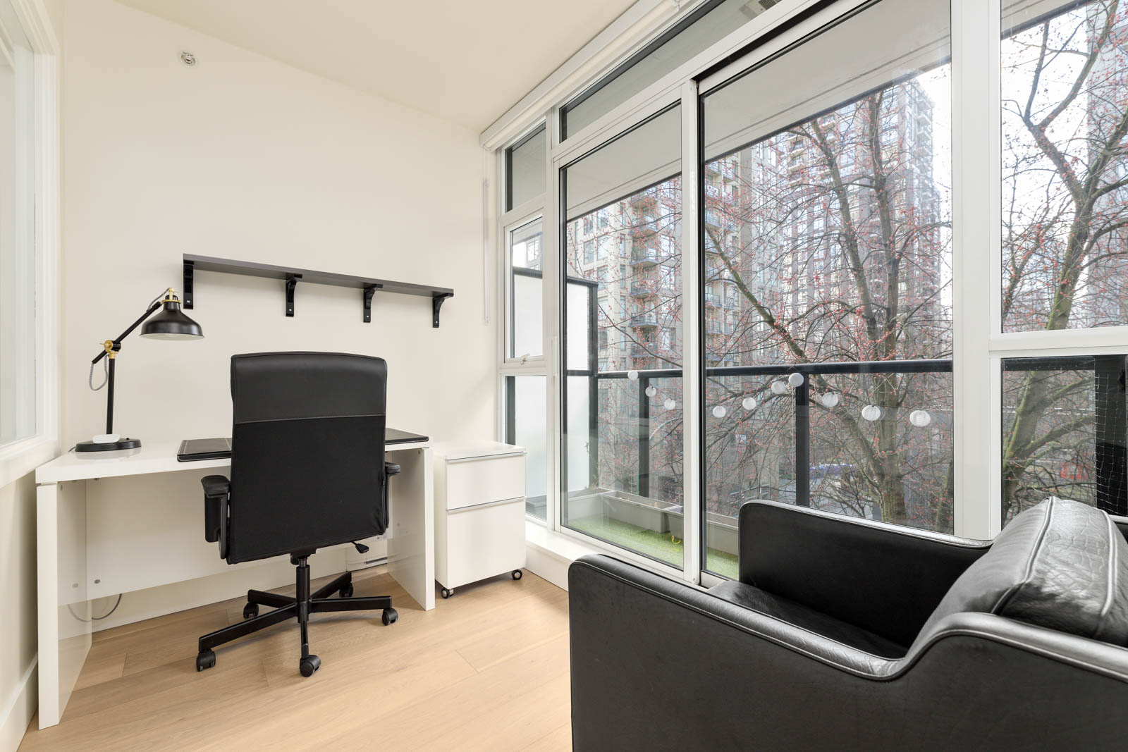 Minimalist home office with a desk, chair, lamp, and shelf, next to large windows overlooking a balcony and city buildings. Black armchair in the foreground.
