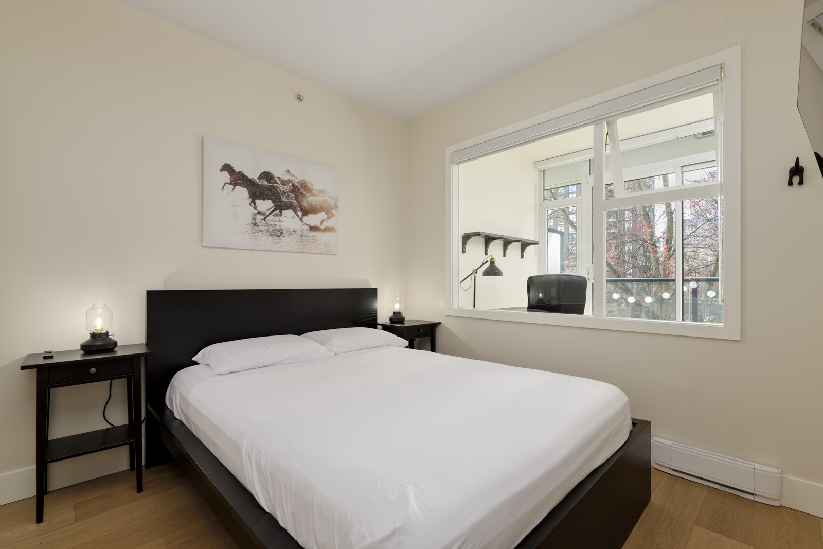 A minimalist bedroom with a black bed frame, white bedding, two black nightstands, lamps, and a black-and-white horse photo. A window shows an adjacent office space with a desk and chair.