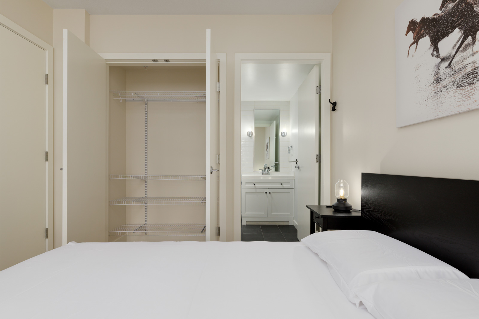 A tidy bedroom with a made bed, open closet with shelves, and a view into a bathroom with a sink and mirror.