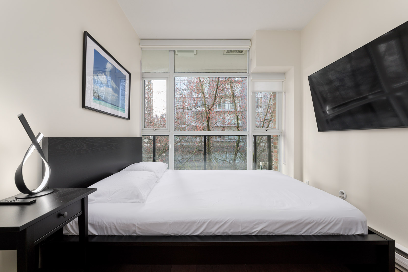 Modern bedroom with a neatly made bed, black headboard, nightstand with a lamp, wall-mounted TV, and large window showing trees outside.