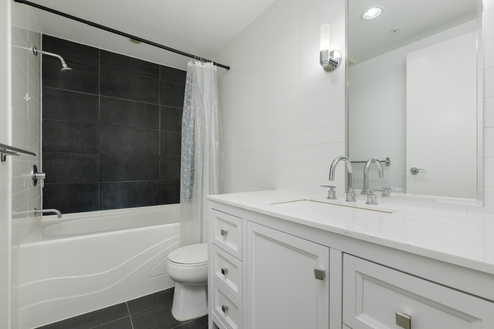 Modern bathroom with a white vanity, two sinks, a large mirror, toilet, bathtub with a white shower curtain, and black and white tiled walls and floor.