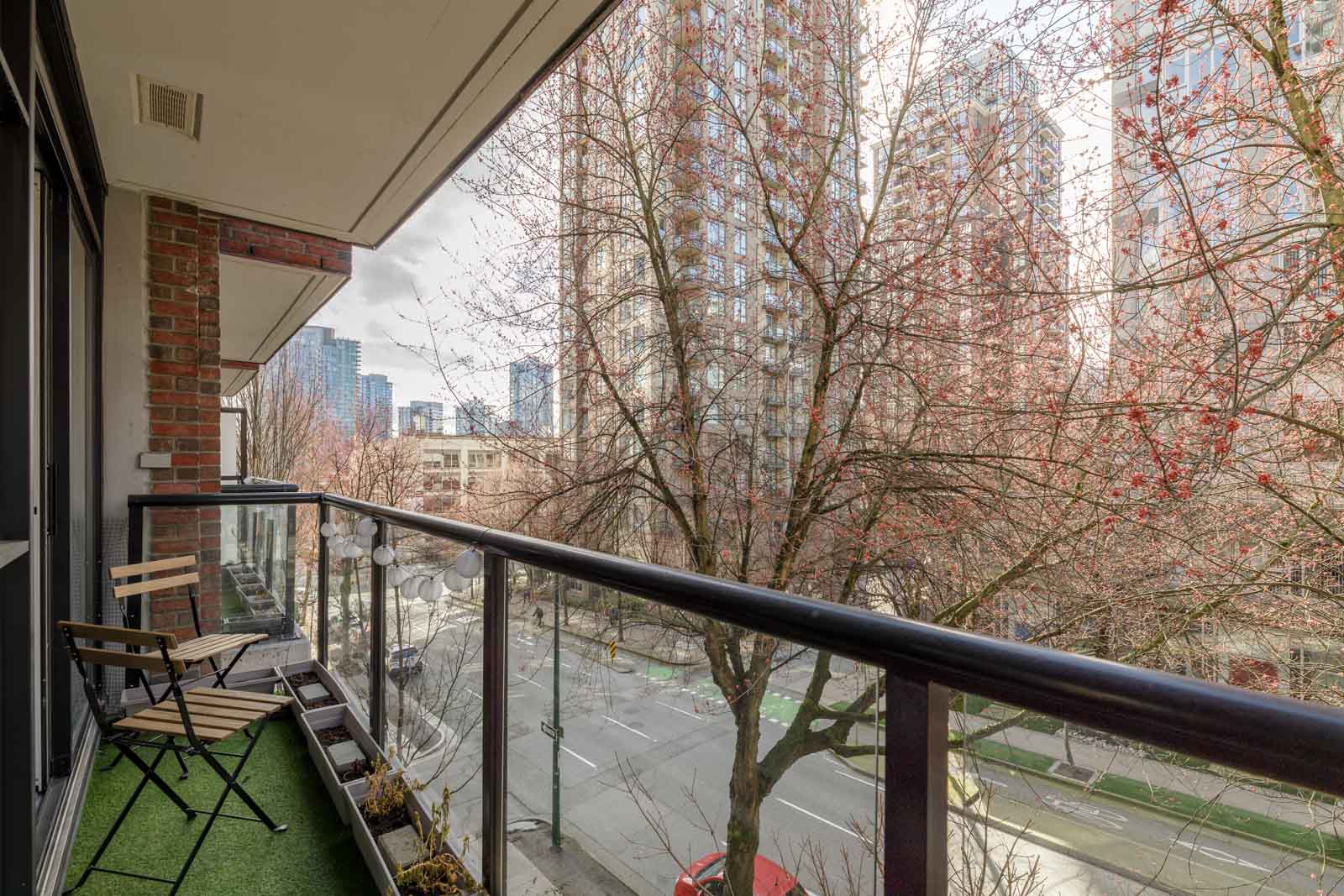 A small balcony with artificial grass and a table and chairs overlooks a street lined with leafless trees and tall apartment buildings.