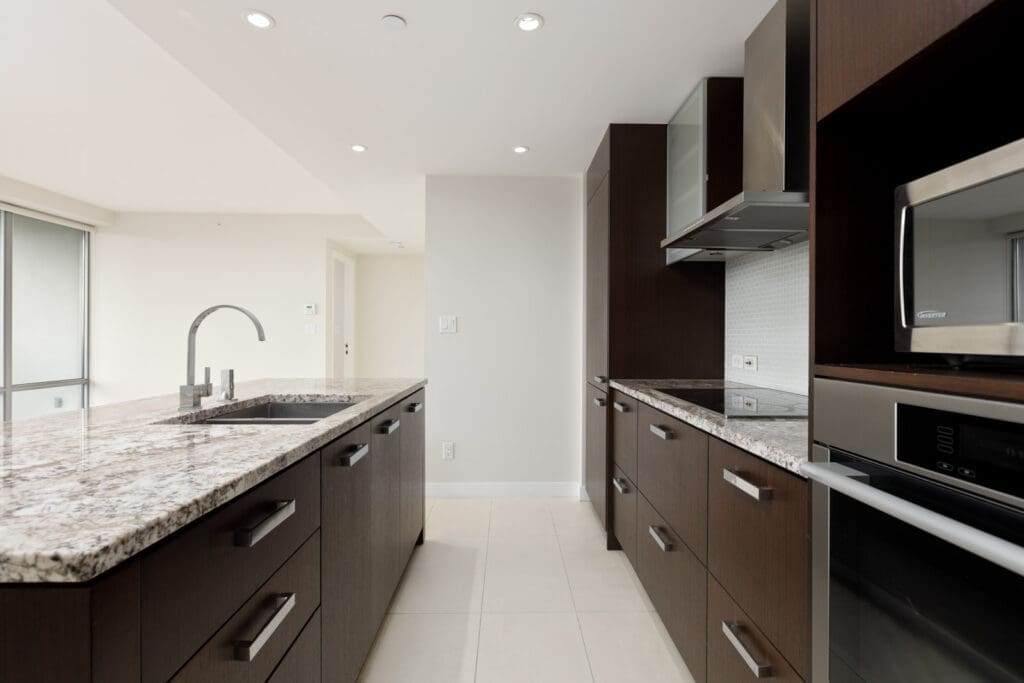 Modern kitchen with dark wood cabinets, granite countertops, stainless steel appliances, and a central island with a sink under recessed lighting.