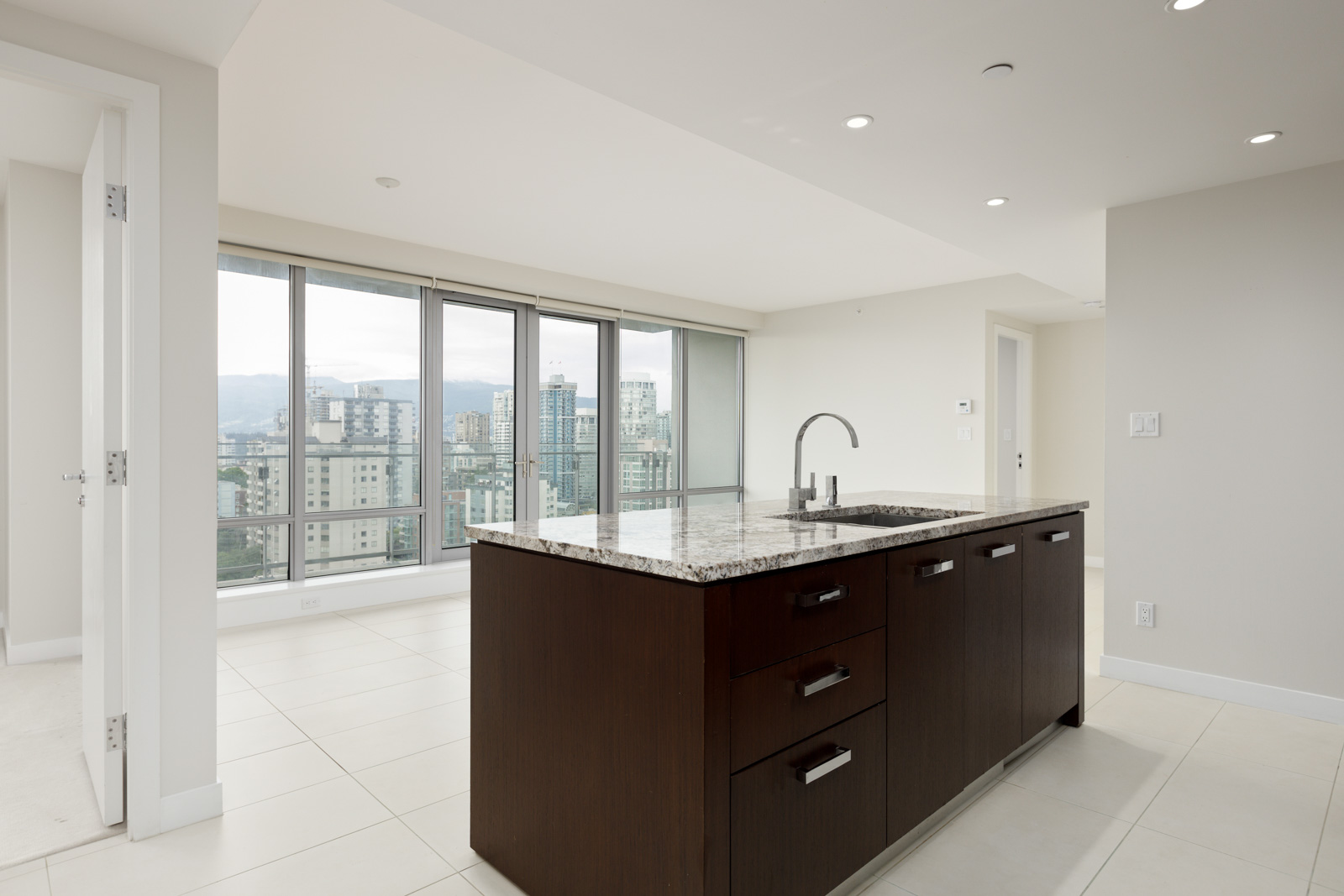 Modern, unfurnished apartment with large windows, city view, neutral walls, and a kitchen island featuring a granite countertop and built-in sink.