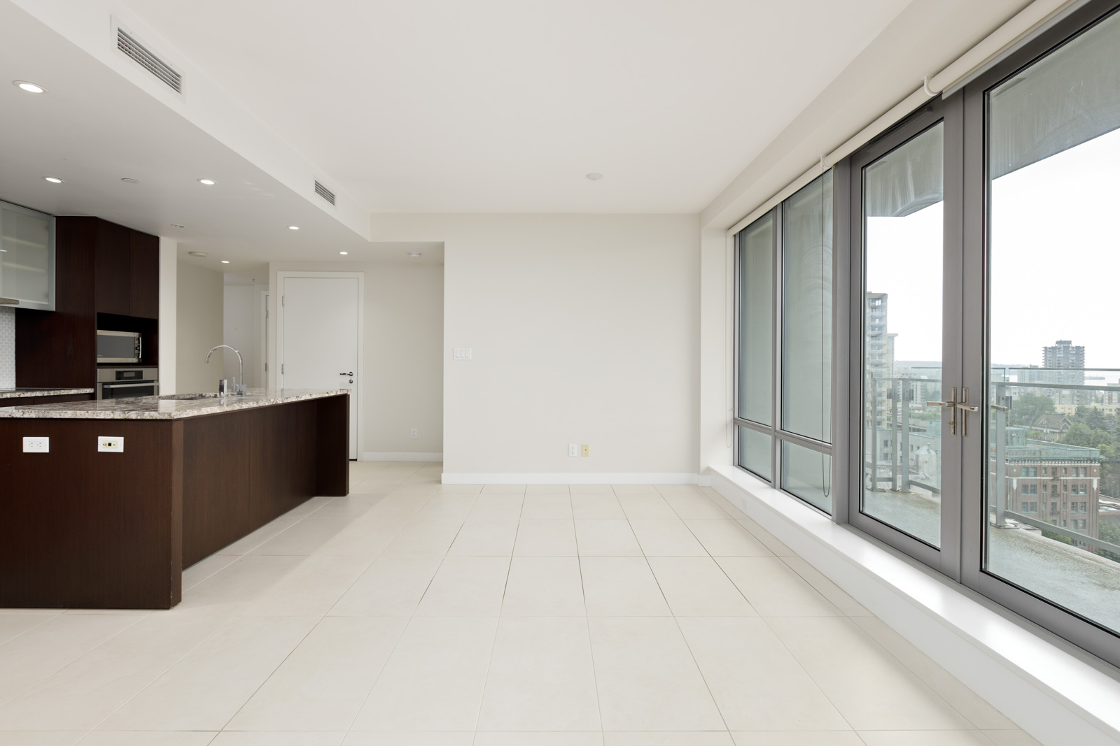 Modern, unfurnished apartment with large windows, tiled floors, and an open kitchen featuring dark wood cabinets and granite countertops, overlooking a cityscape.
