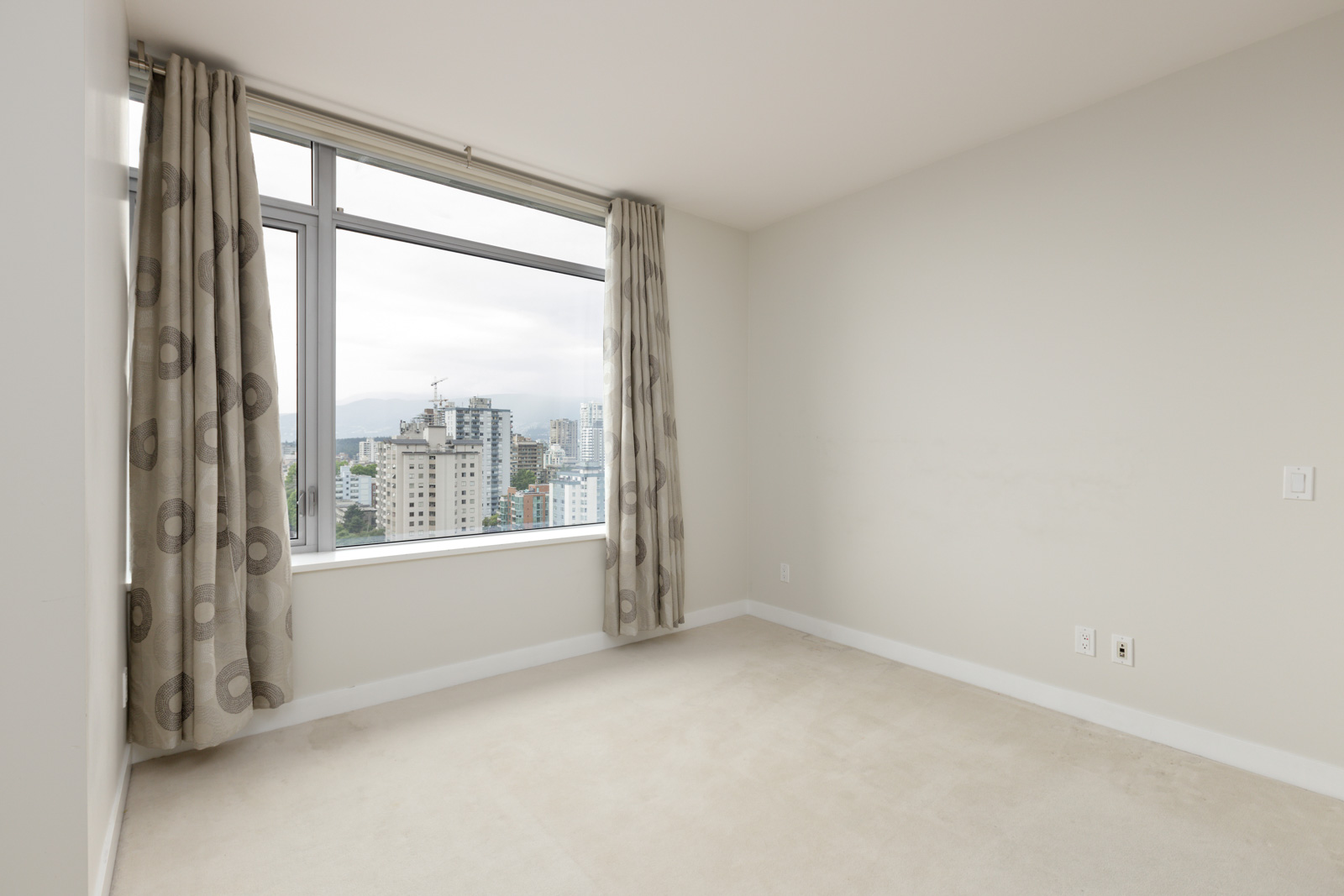 Unfurnished room with beige carpet, pale walls, and a large window with patterned curtains overlooking a cityscape.