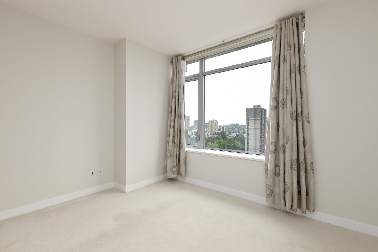 Empty room with beige carpet, light-colored walls, a large window with patterned curtains, and a cityscape view outside.