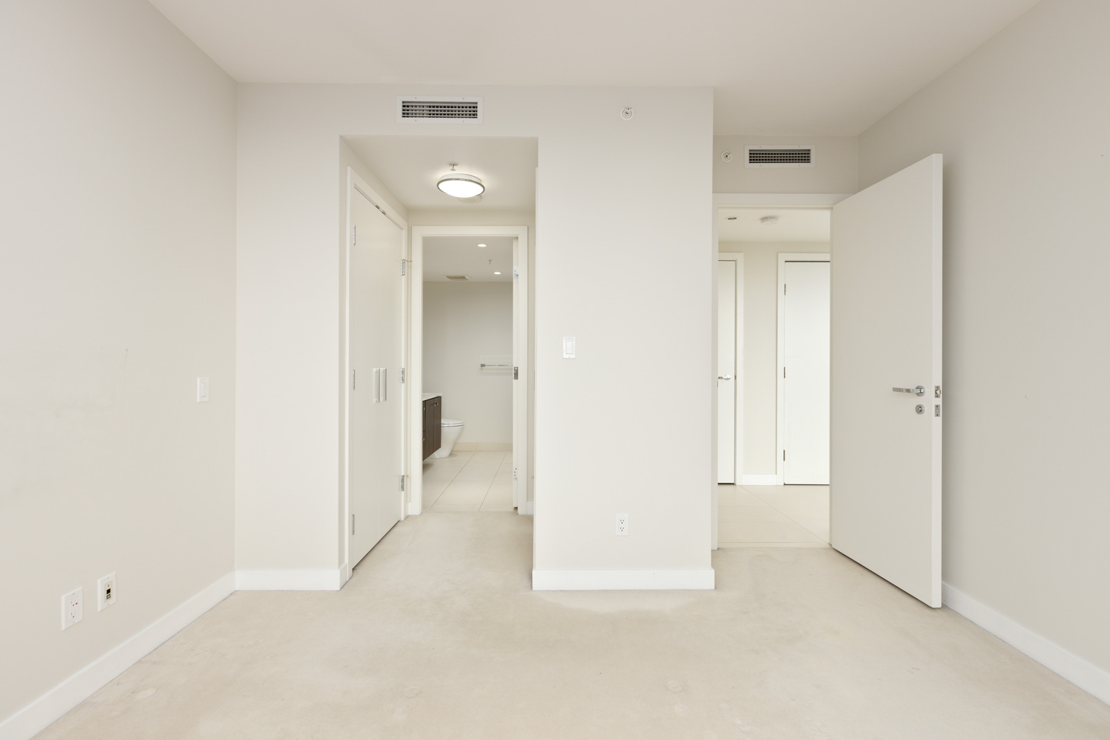 Empty, unfurnished room with beige carpet, white walls, and three open doors leading to a bathroom, closet, and hallway. Ceiling vent and light fixture are visible.