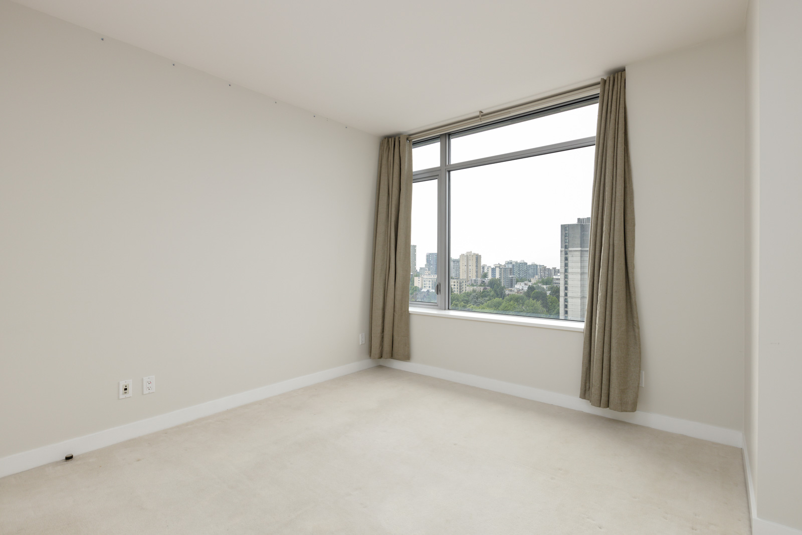 Empty room with beige carpet, white walls, two electrical outlets, and a large window with beige curtains overlooking a cityscape.