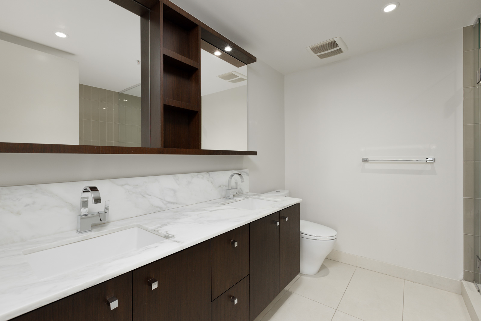 Modern bathroom with a marble countertop, dual sinks, dark wood cabinets, a large mirror, toilet, towel rack, and neutral tile flooring.