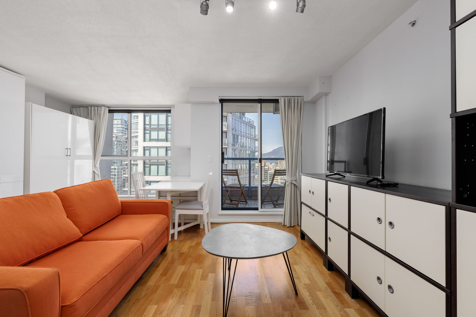 A modern apartment living room with an orange sofa, round coffee table, TV on a cabinet, dining table by the window, and balcony view of city buildings.