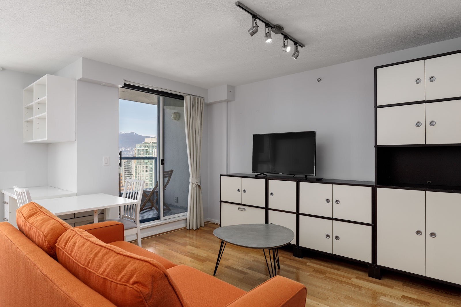 Modern living room with an orange sofa, white table with two chairs, TV on a black and white cabinet, and a sliding door to a balcony with city views.