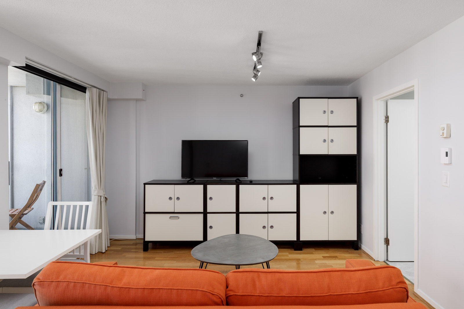 A modern living room with an orange sofa, a round coffee table, a black-and-white TV stand with a flat-screen TV, and a balcony visible through a sliding glass door.