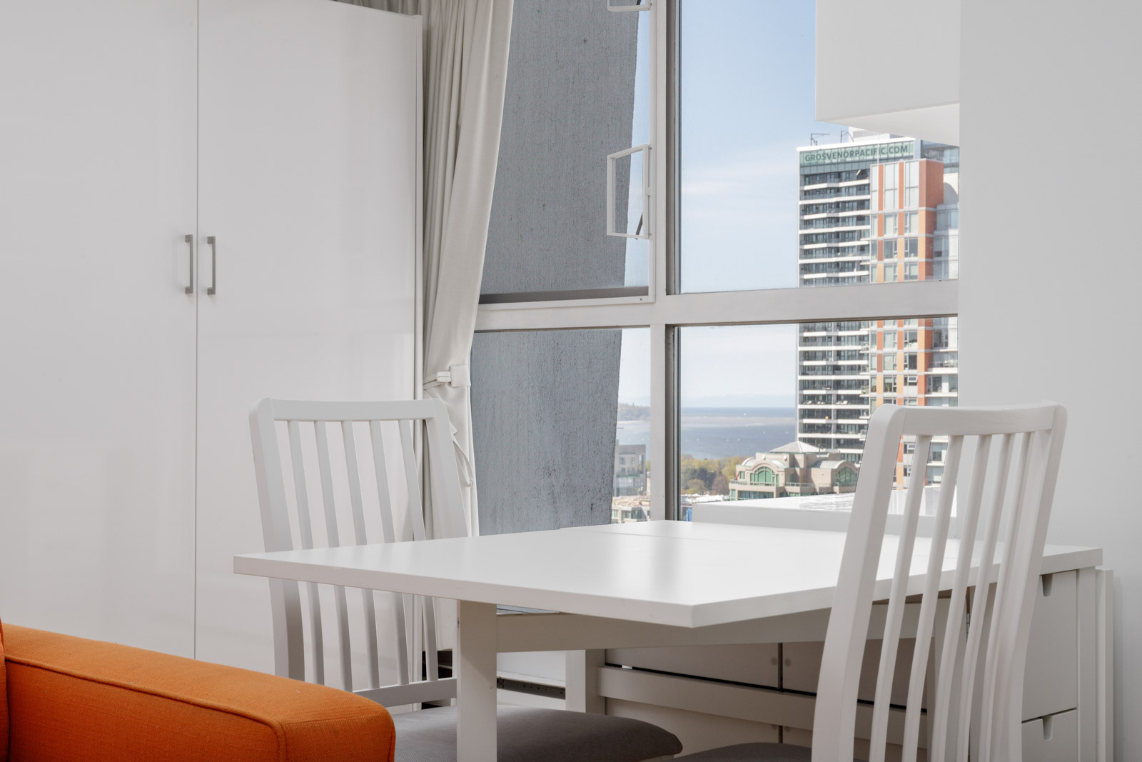 A small dining area with a white table and two white chairs next to a window, overlooking city buildings and a distant view of water.