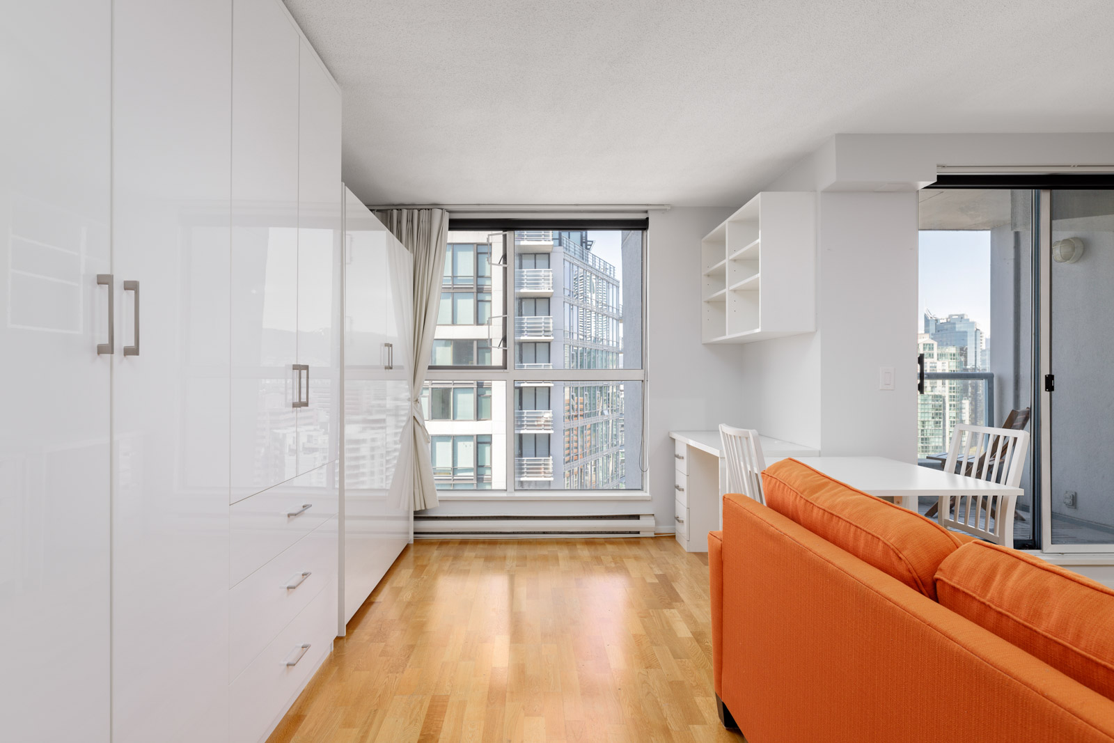 Bright modern apartment interior with wood flooring, white cabinetry, a small desk, orange sofa, and large windows overlooking city buildings and a balcony.