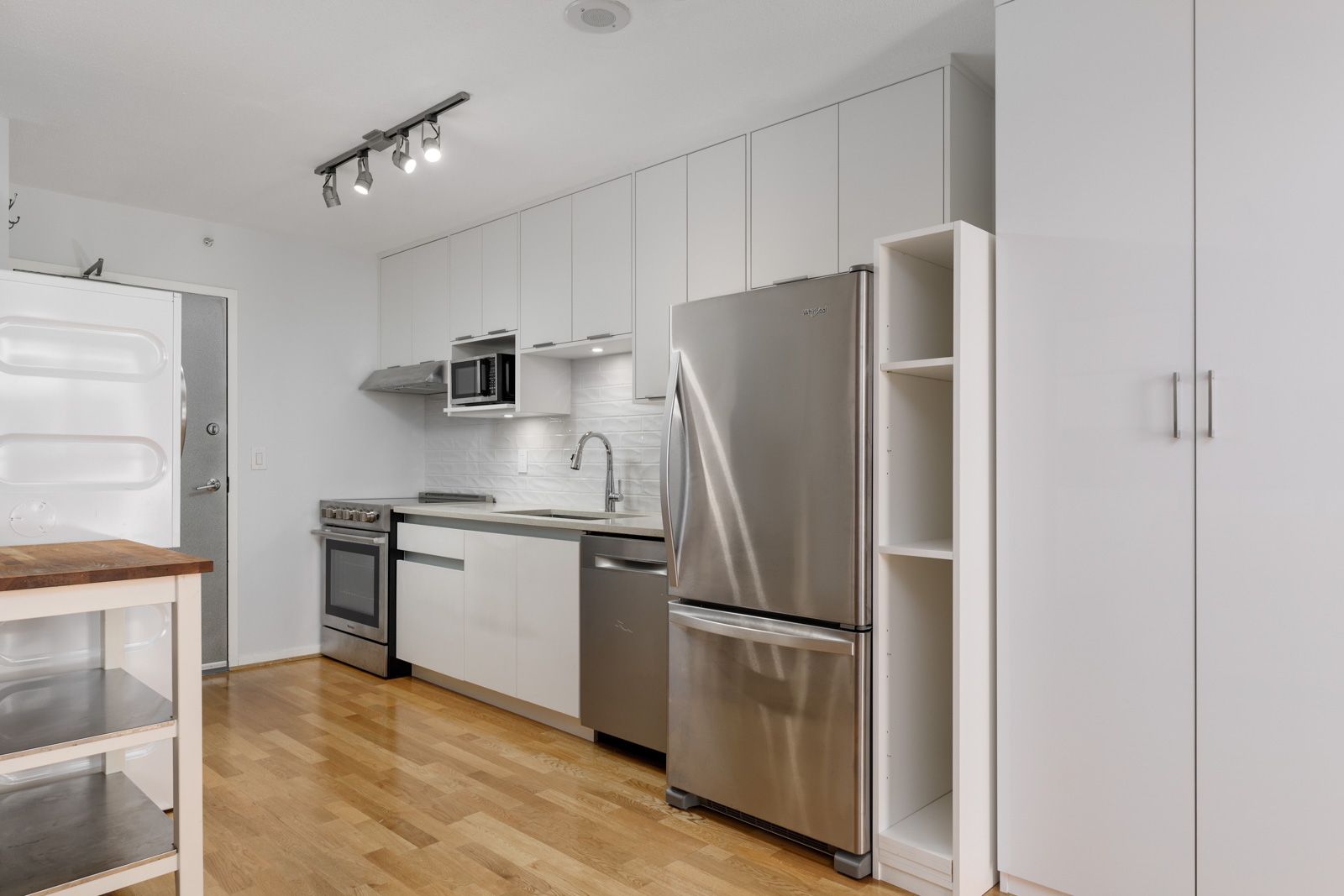 Modern kitchen with white cabinets, stainless steel appliances, wood flooring, track lighting, and a small open shelf next to the refrigerator.