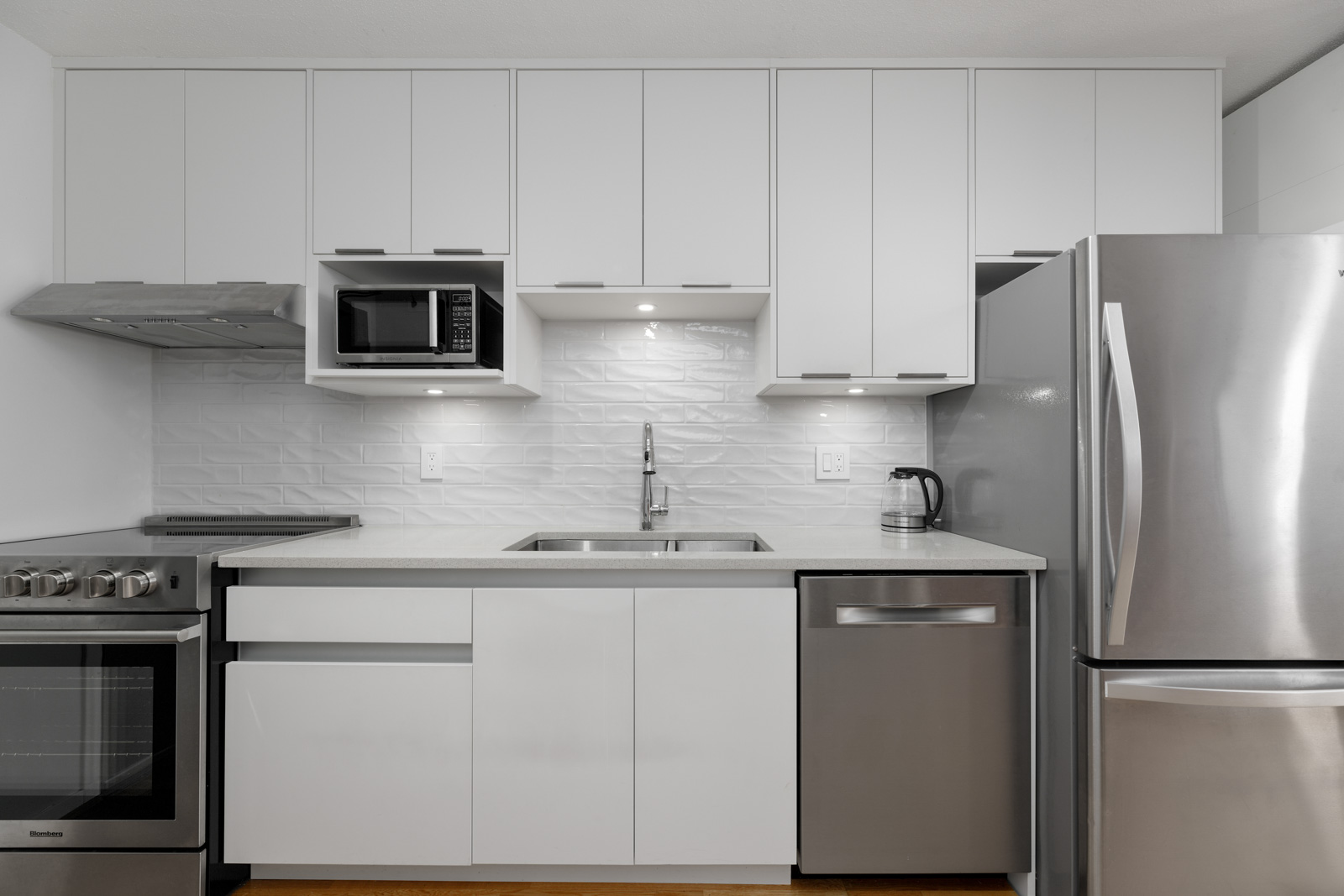 Modern kitchen with white cabinets, stainless steel appliances, a sink under a central light, and a white textured backsplash.