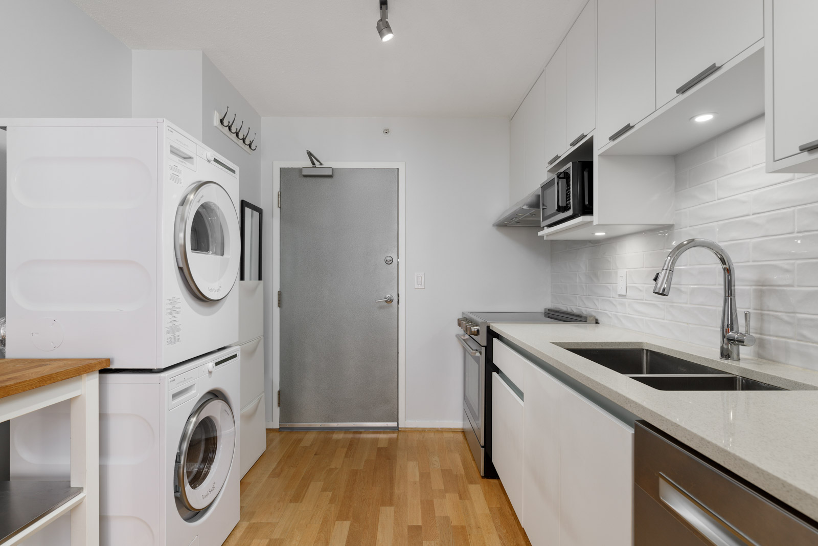 A modern kitchen with light wood flooring, white cabinets, a stacked washer and dryer, stainless steel appliances, and a silver door.