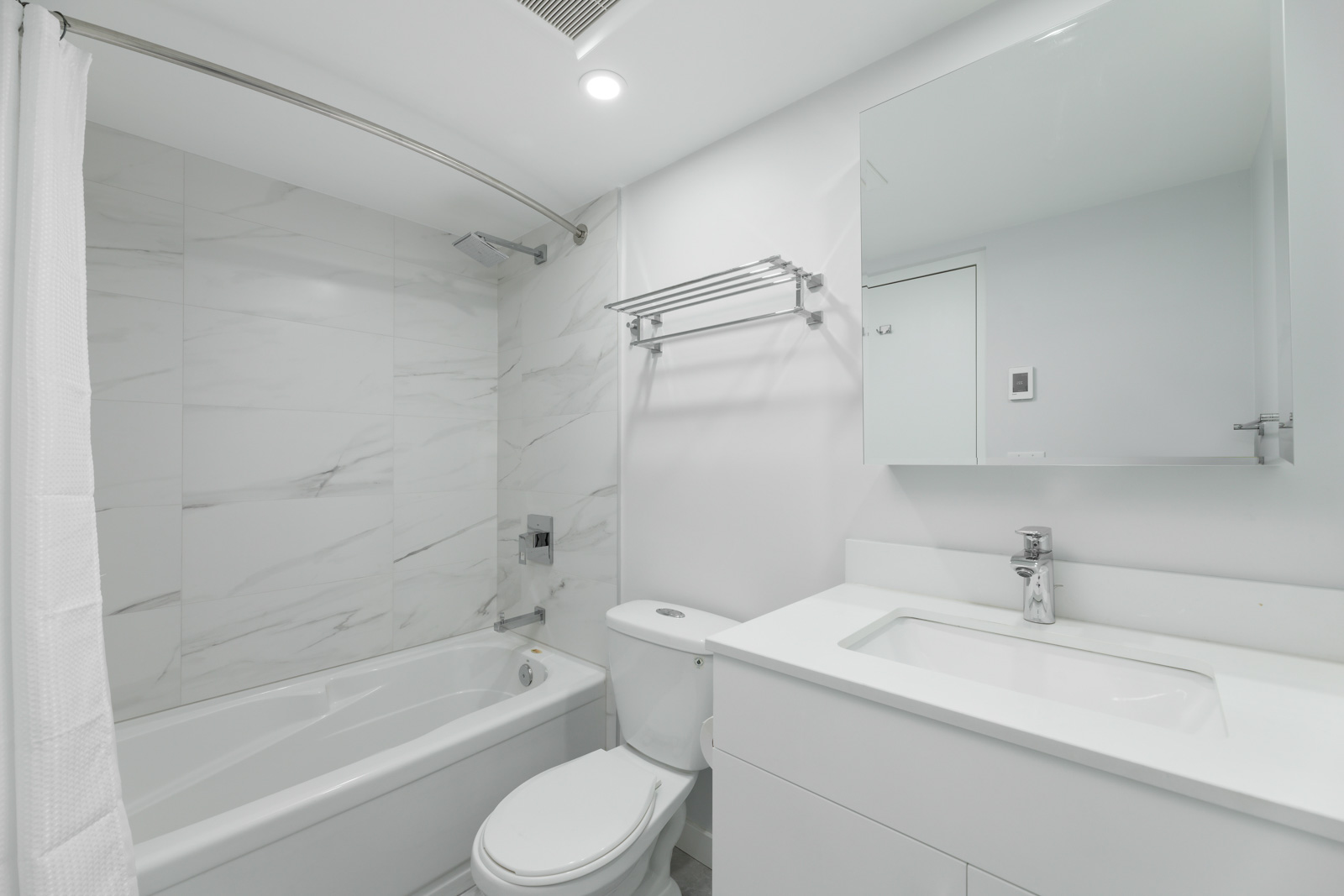 Modern white bathroom with a bathtub, toilet, wall-mounted towel rack, and a sink with a mirrored cabinet above. Marble tiles line the shower wall.