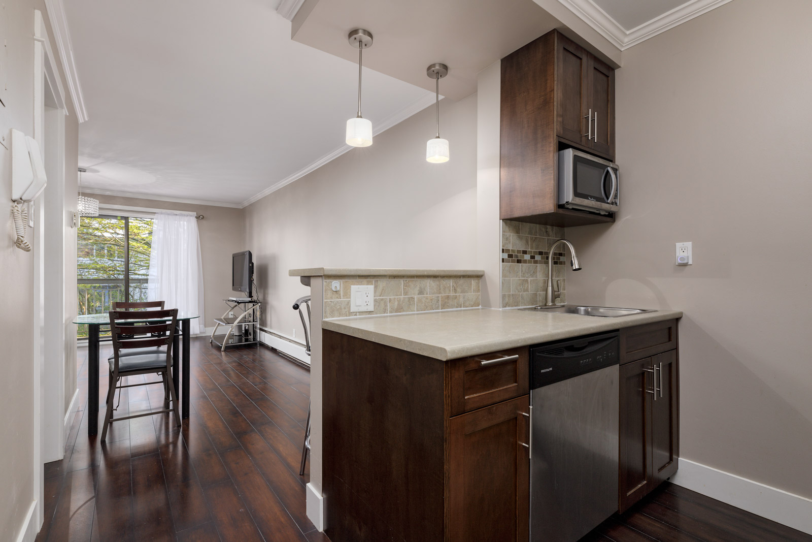 Modern apartment kitchen with dark wood cabinets, stainless steel appliances, and a small dining area by a window with sheer curtains.