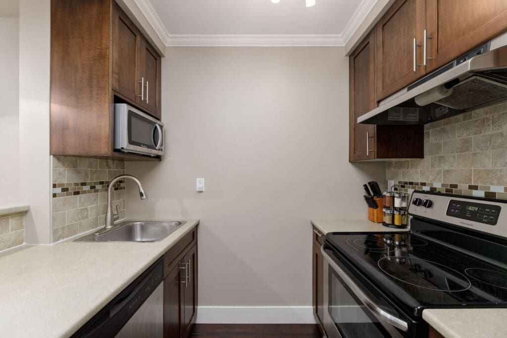 A modern kitchen with dark wood cabinets, beige countertops, a stainless steel sink, electric stove, microwave, and tiled backsplash.