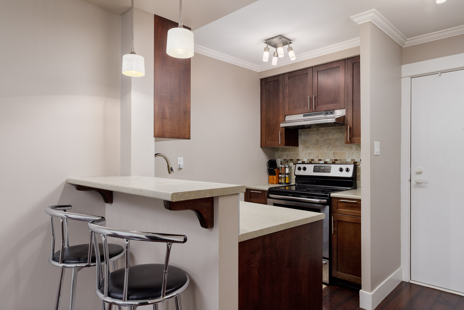 Modern kitchen with dark wood cabinets, stainless steel appliances, a breakfast bar with two metal stools, and neutral-colored walls.