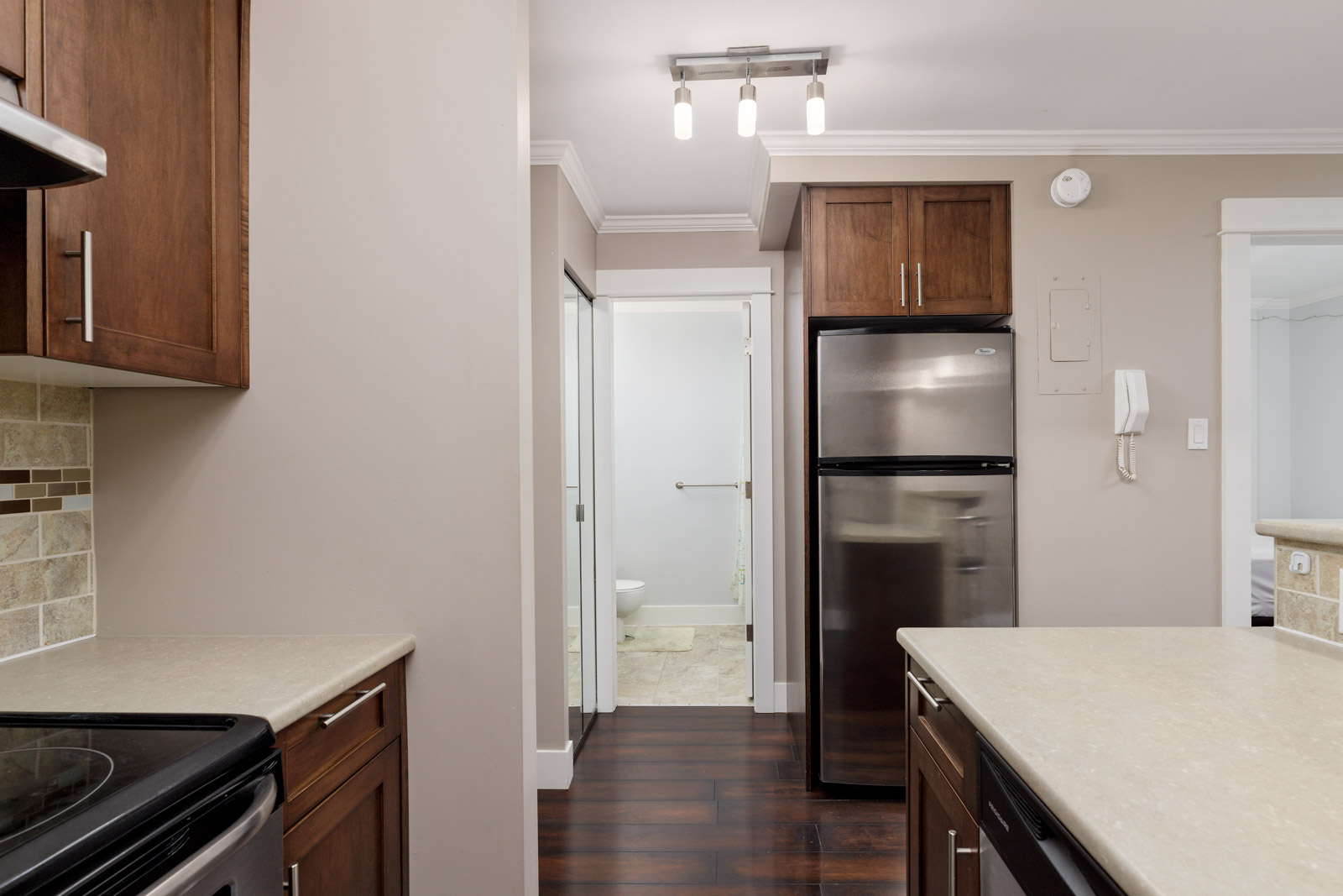 View of a modern kitchen with dark cabinets, stainless steel appliances, beige countertops, and a glimpse of a bathroom through a doorway in the background.
