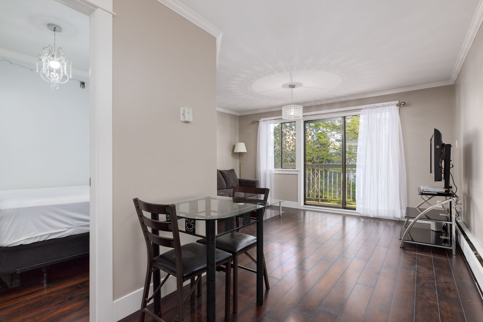 A modern apartment living room with dark hardwood floors, a glass dining table with chairs, a TV on a stand, and large windows with sheer curtains leading to a balcony.