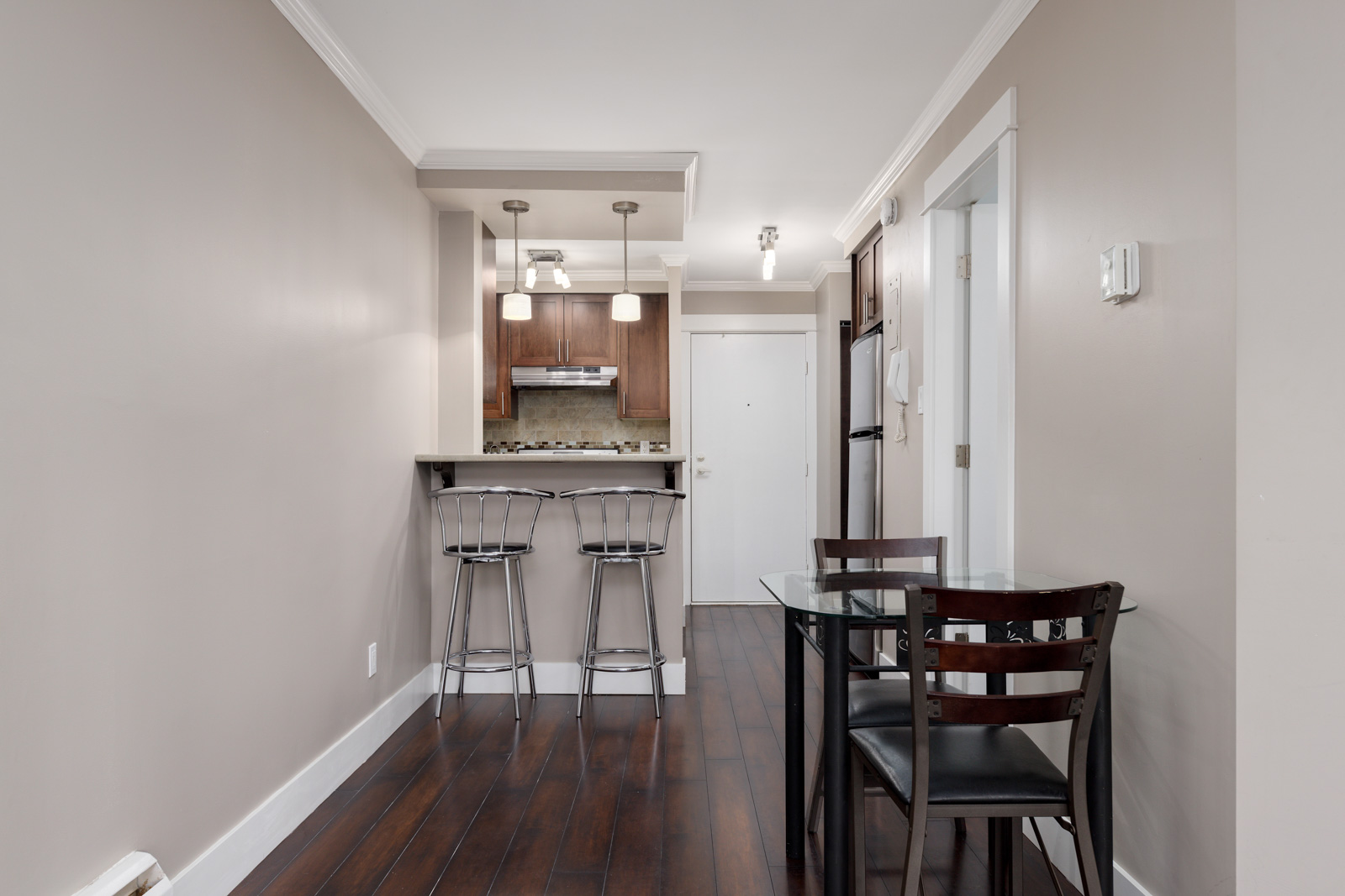 Modern kitchen and dining area with dark wood flooring, a small glass dining table with three chairs, and a breakfast bar with two stools under pendant lights.