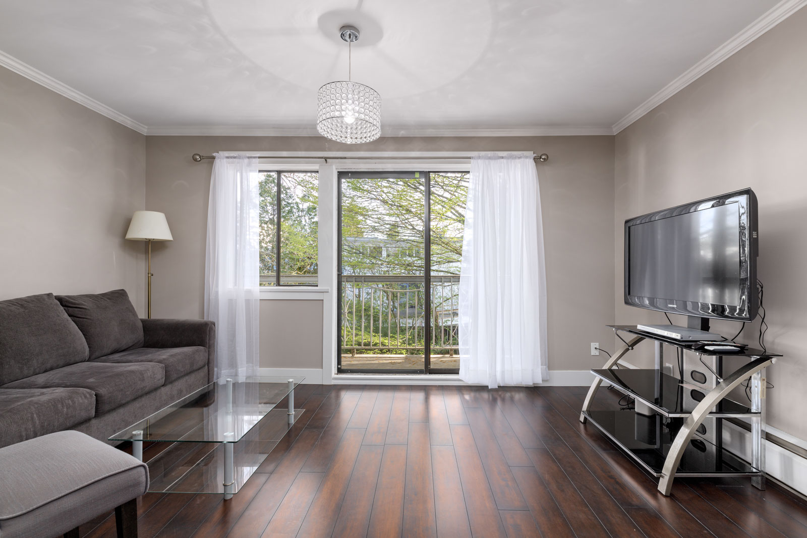 Modern living room with a gray sofa, glass coffee table, TV on a stand, floor lamp, hardwood floors, and sliding glass doors with white curtains leading to a balcony.