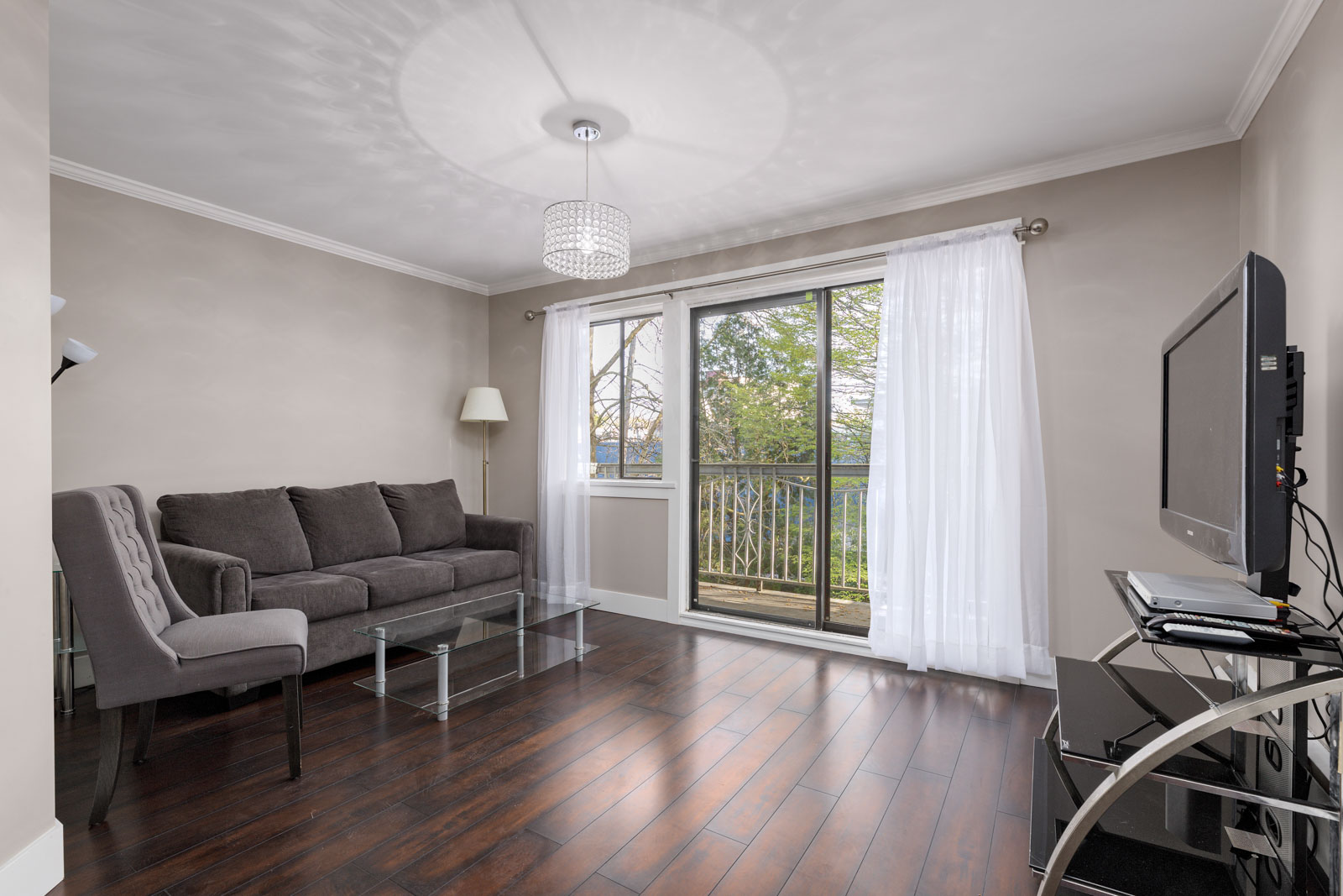 Modern living room with a gray sofa, armchair, glass coffee table, TV on a stand, and sliding glass doors leading to a balcony with a view of trees.