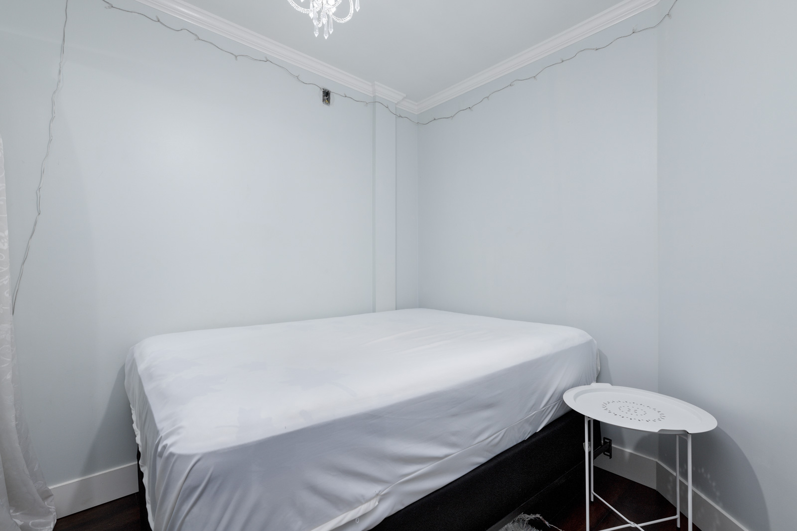 A small room with a neatly made bed, light blue walls, a white round side table, dark floor, and a ceiling light fixture.
