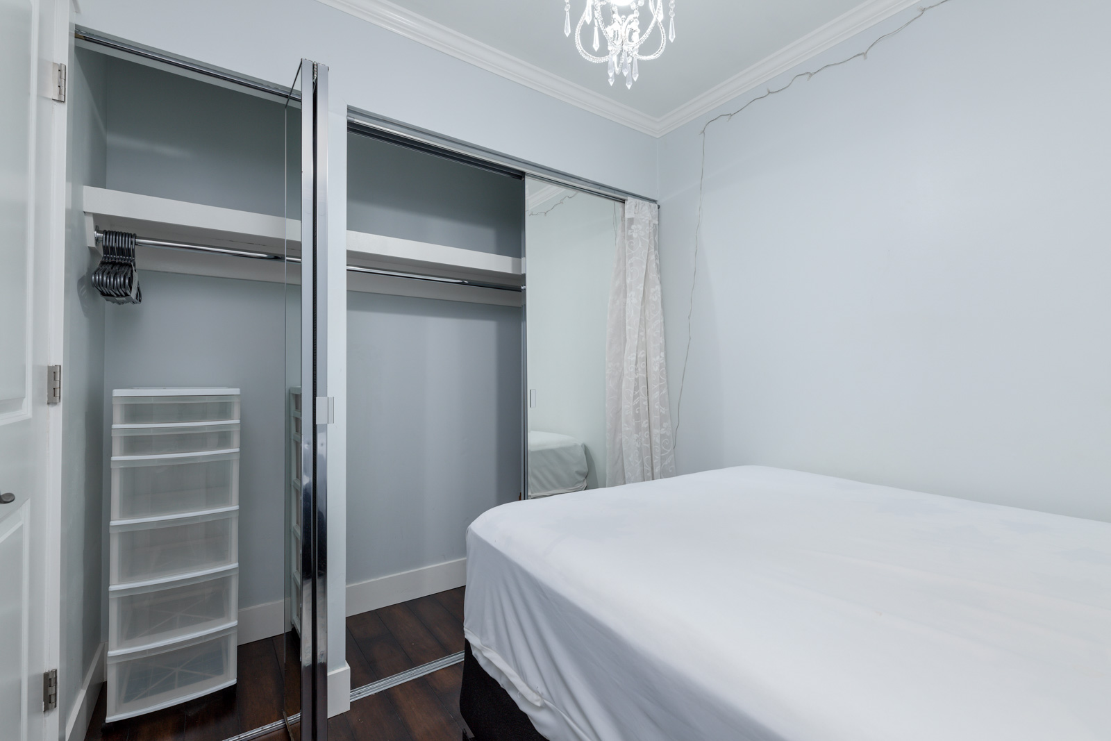A small bedroom with a neatly made bed, mirrored sliding closet doors, plastic storage drawers, hangers, and a chandelier on the ceiling.