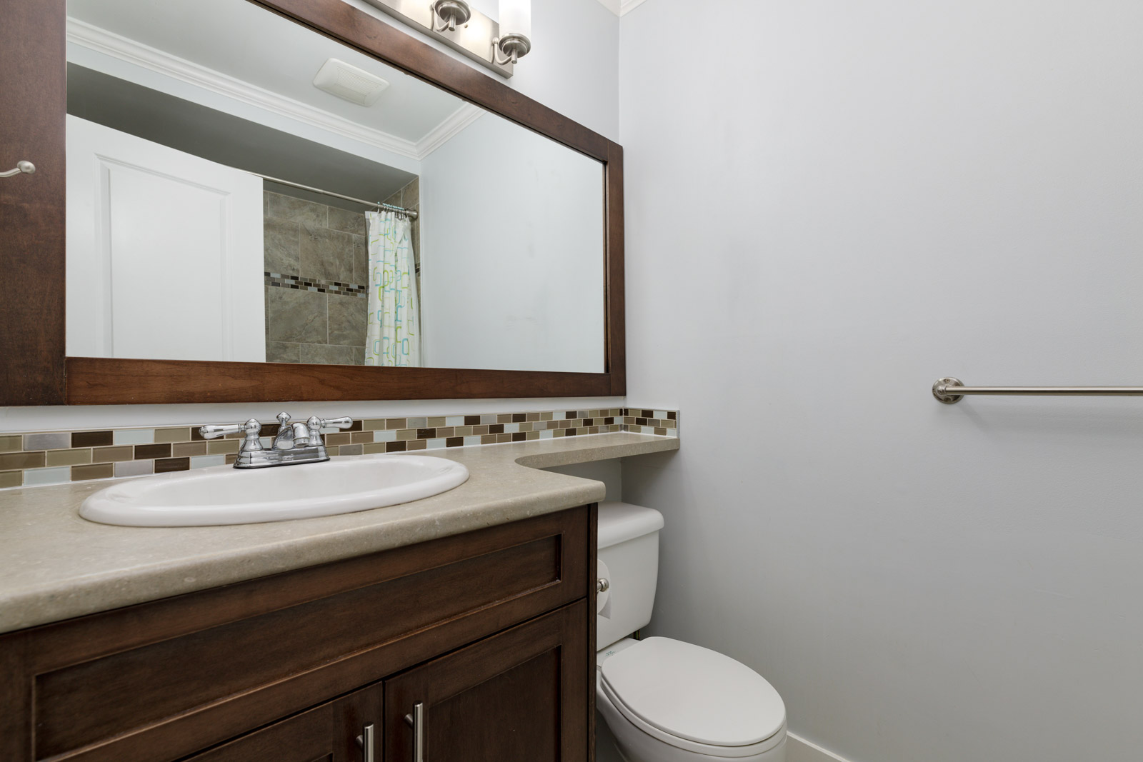 A bathroom with a single sink vanity, large mirror, toilet, towel bar, and a shower with a patterned curtain in the background.