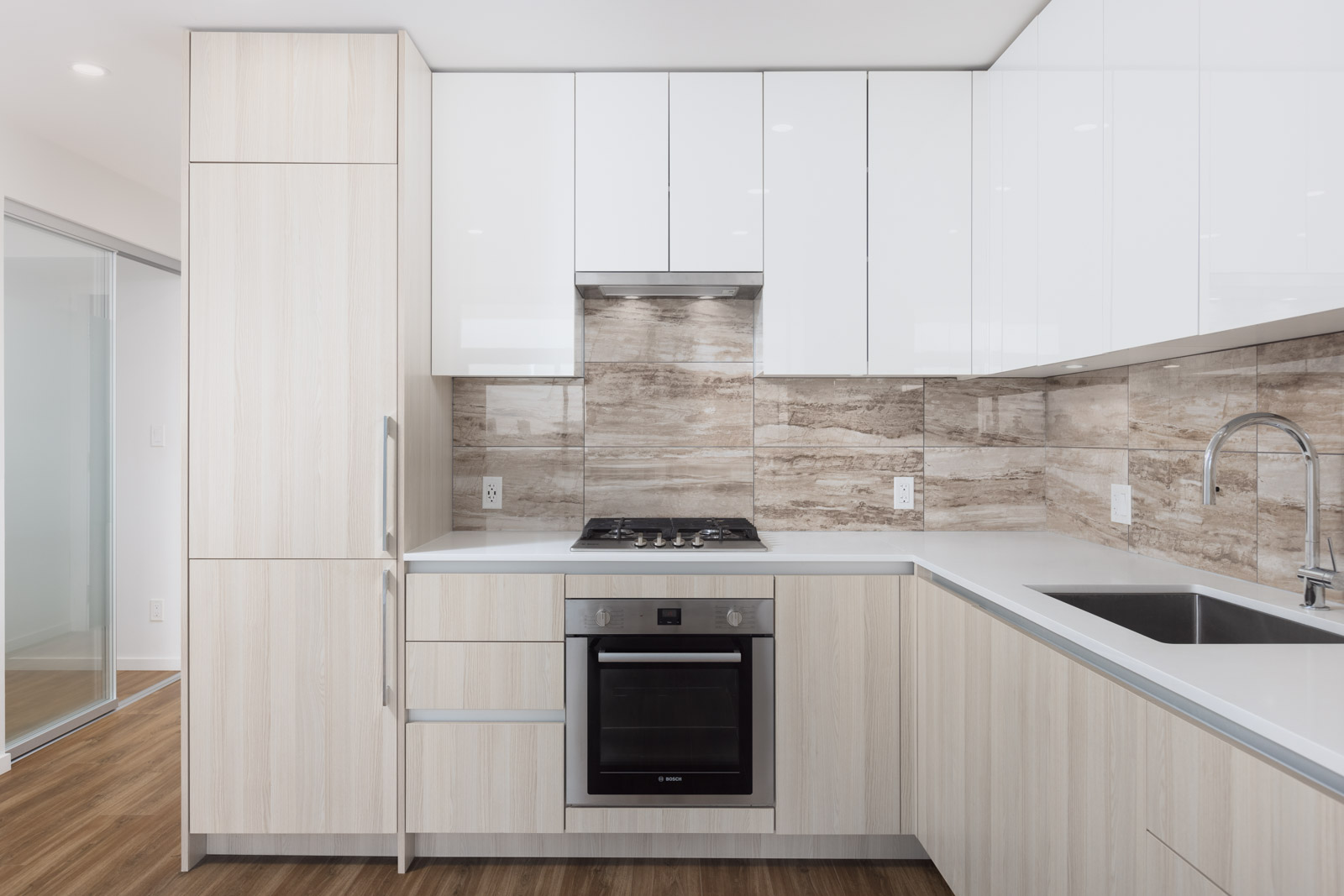 Modern kitchen with light wood cabinets, white countertops, built-in oven, gas cooktop, stainless steel sink, and beige tile backsplash.