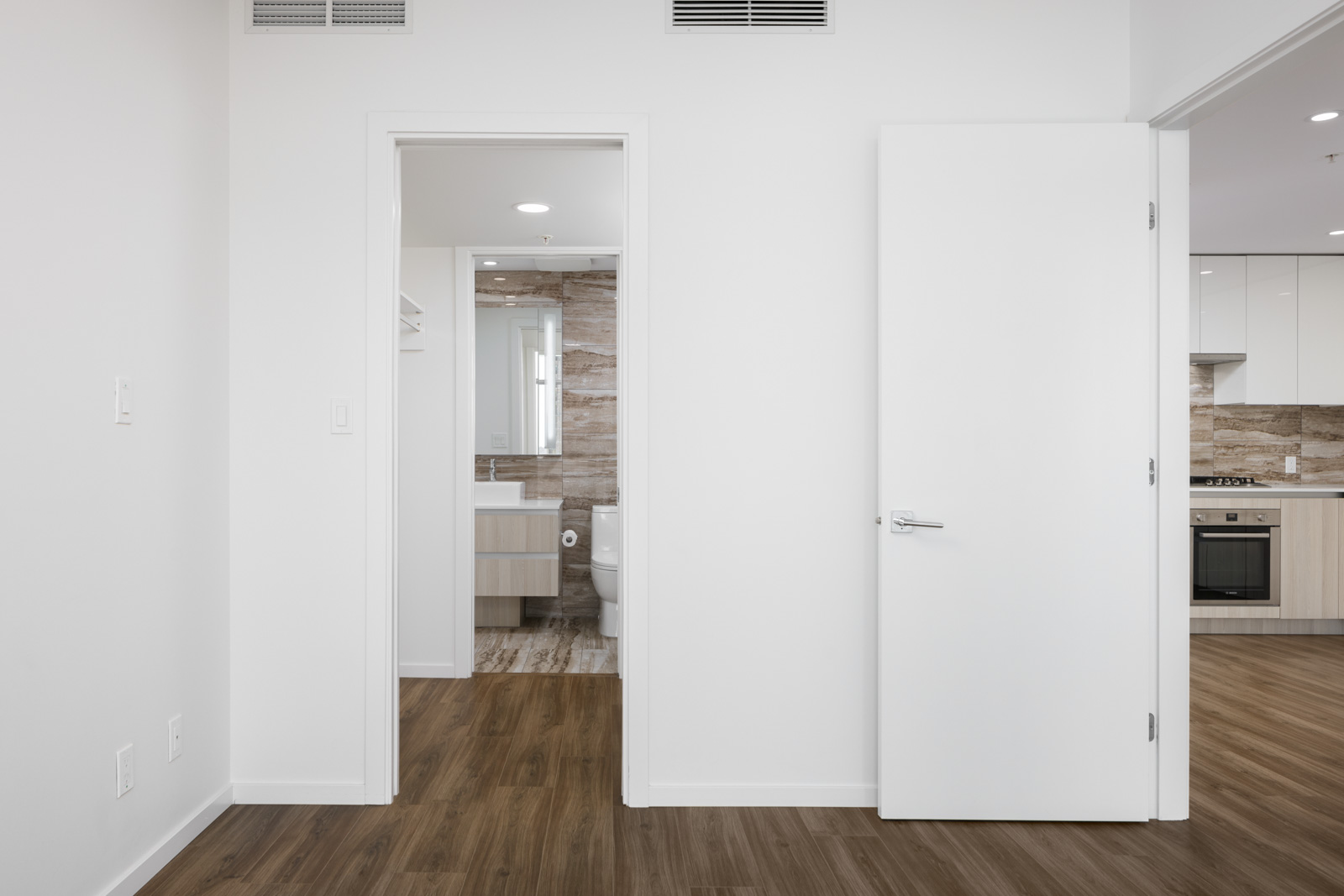 A modern, unfurnished apartment room with wood floors, white walls, an open door, visible kitchen, and a bathroom with tile accents seen through a doorway.