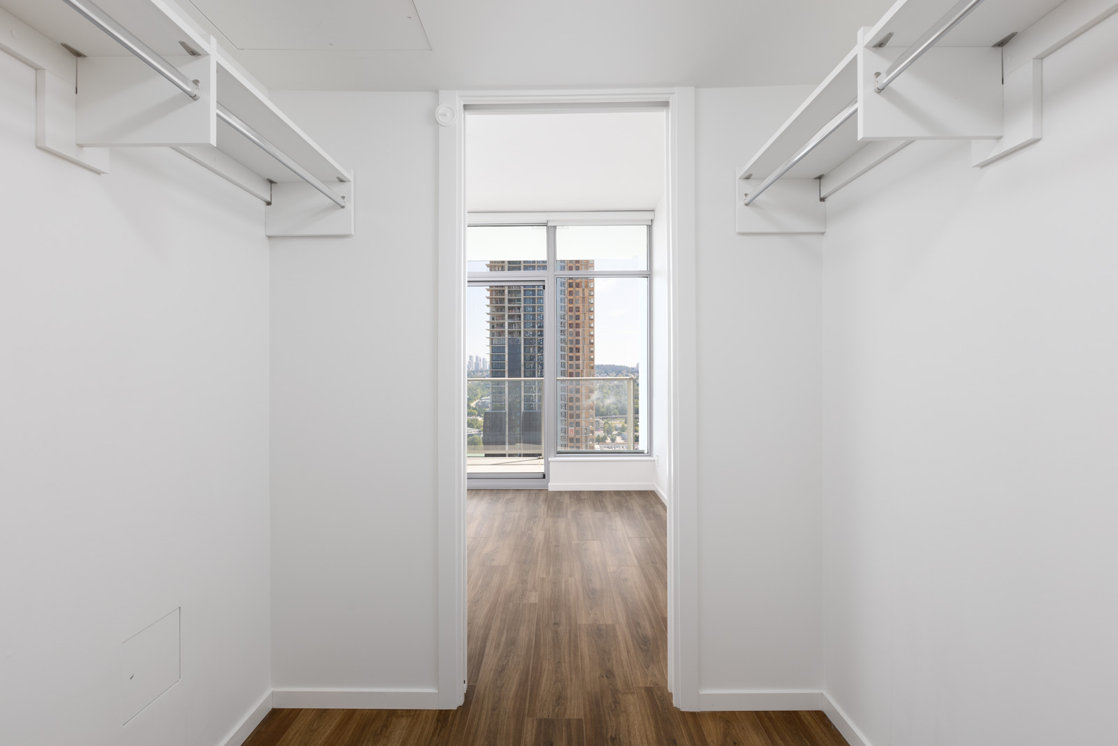 Empty walk-in closet with white walls and shelving, leading to a room with large windows and a view of high-rise buildings and a cityscape.