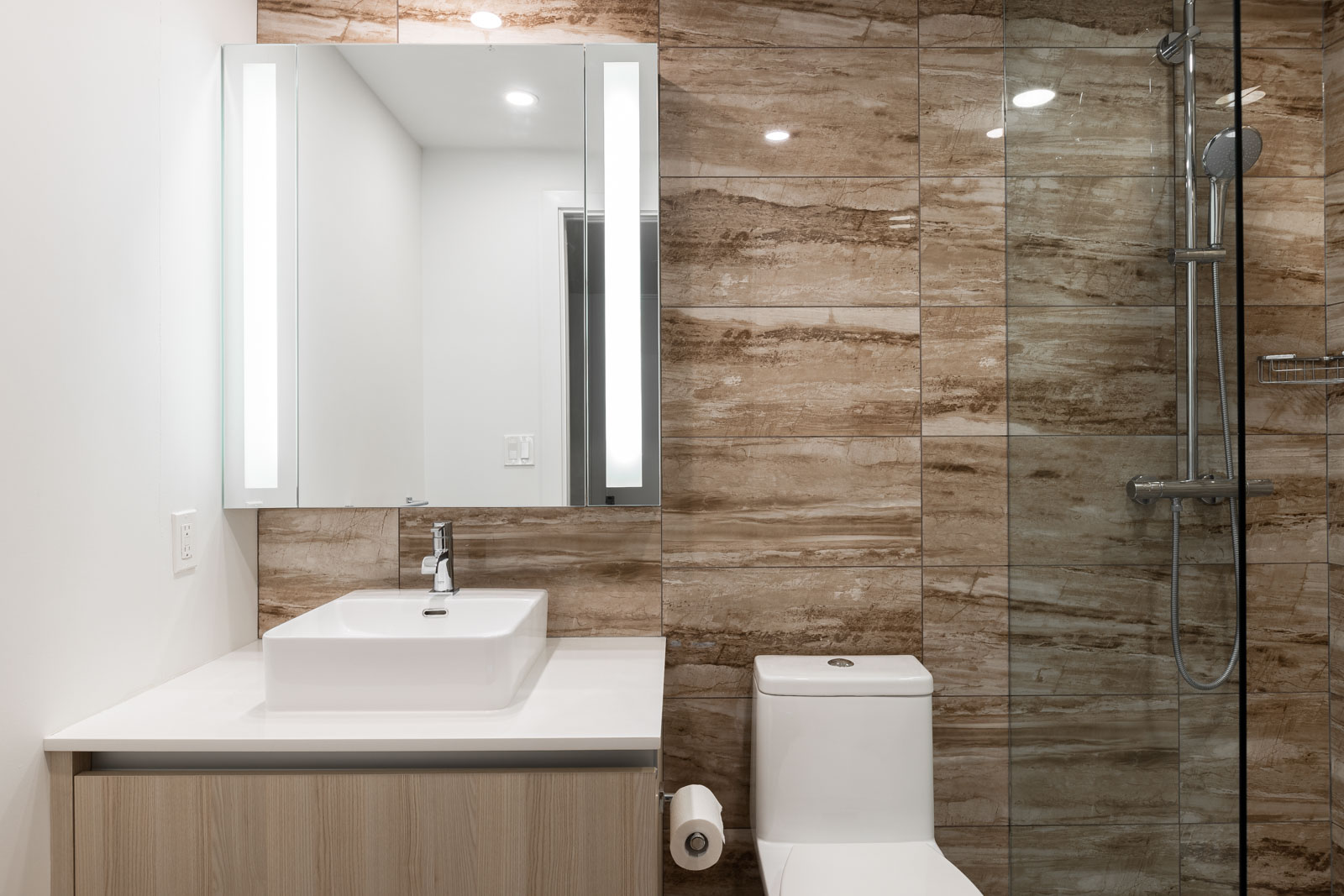 Modern bathroom with a square sink, wall-mounted mirror, glass shower, and beige marble tiles on the wall behind the toilet and shower area.