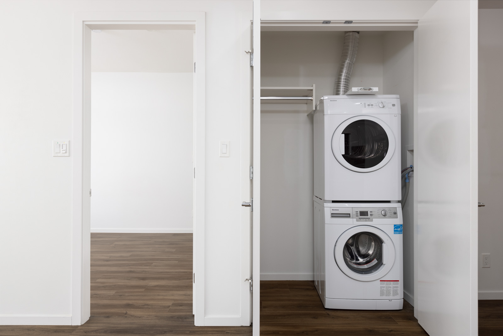 A stacked washer and dryer are installed in a closet with white doors, next to an empty room with wood flooring and white walls.